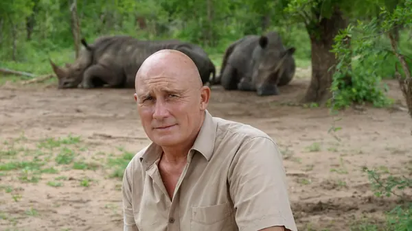 Patrick en chemise beige, deux rhinocéros au repos en arrière-plan